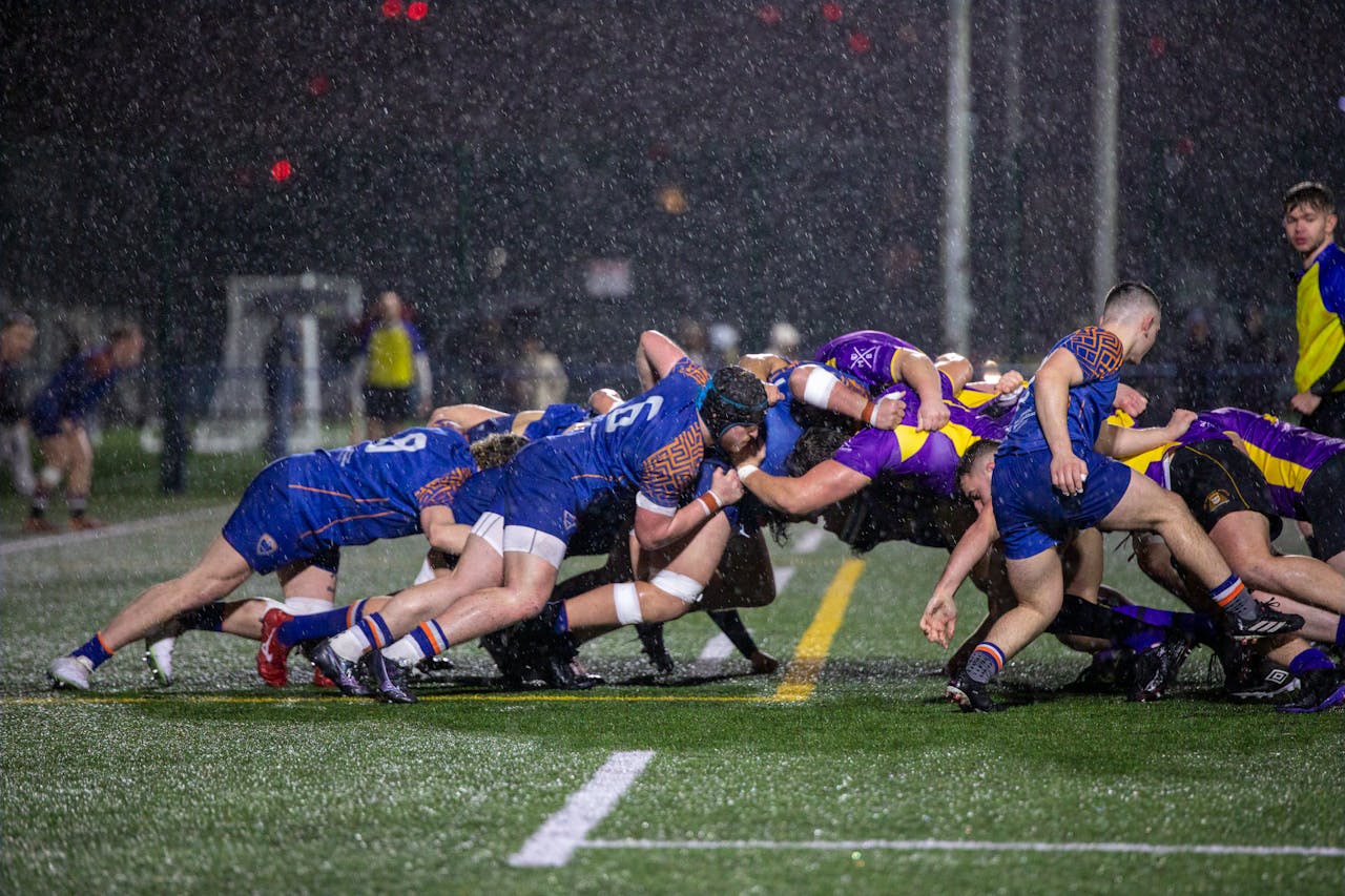 about-img Dramatic rugby scrum at night in Manchester rain, showcasing teamwork and athleticism.